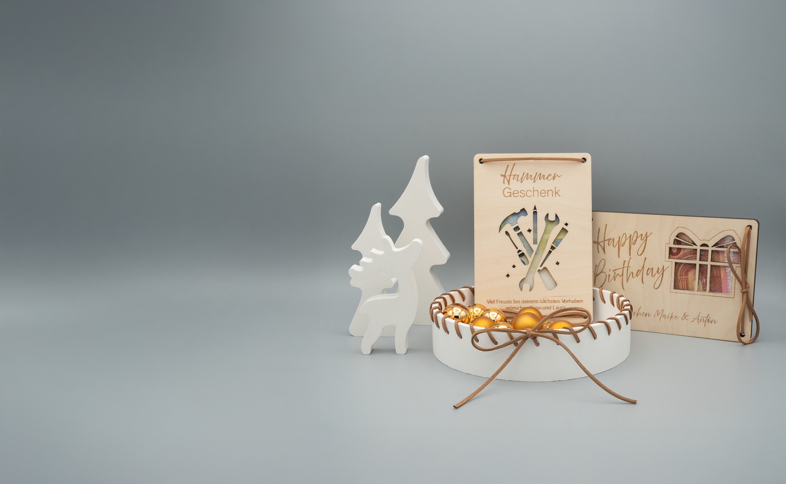 Stimmungsvolle Deko-Szene mit weißem Rentier und Weihnachtsbaum als Figuren, einer weißen Schale mit braunem Kunstlederband verziert und diversen goldenen Weihnachtskugeln. Dahinter stehen zwei Holz-Geschenkkarten mit Gravuren, eine mit dem Schriftzug ‘Hammer Geschenk’ und Werkzeugmotiven, die andere mit ‘Happy Birthday’ und einem Geschenkmotiv.