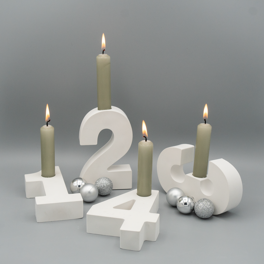 Vier weiße Adventszahlen 1 bis 4 aus Gießpulver dienen als Kerzenhalter für graugrüne Stabkerzen. Um die Zahlen sind silberne Weihnachtskugeln und Glitzerkugeln dekorativ arrangiert. Alle vier Kerzen brennen.