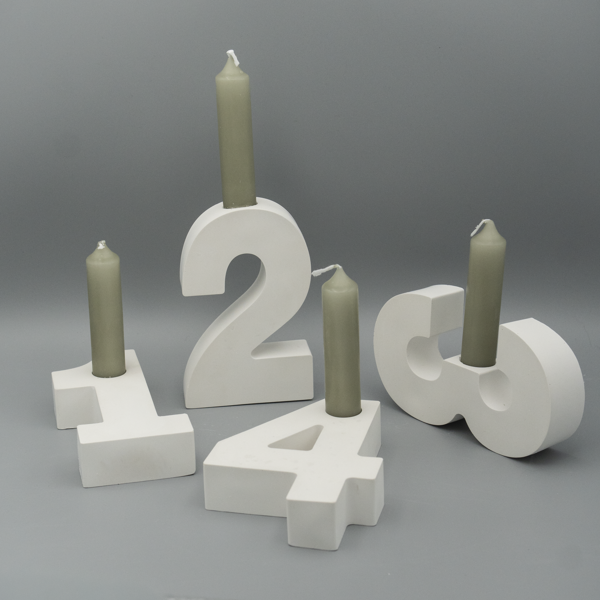Vier Adventszahlen 1 bis 4 aus weißem Gießpulver dienen als Kerzenhalter. In jeder Zahl steckt eine graugrüne Stabkerze, die noch nicht angezündet ist. Die Zahlen sind dekorativ vor einem grauen Hintergrund arrangiert.