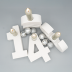 Vier weiße Kerzenhalter in Form der Zahlen 1, 2, 3 und 4 sind aus der Vogelperspektive fotografiert. In jeder Zahl steckt eine brennende Stabkerze. Zwischen den Zahlen liegen silberne und glitzernde Weihnachtskugeln.