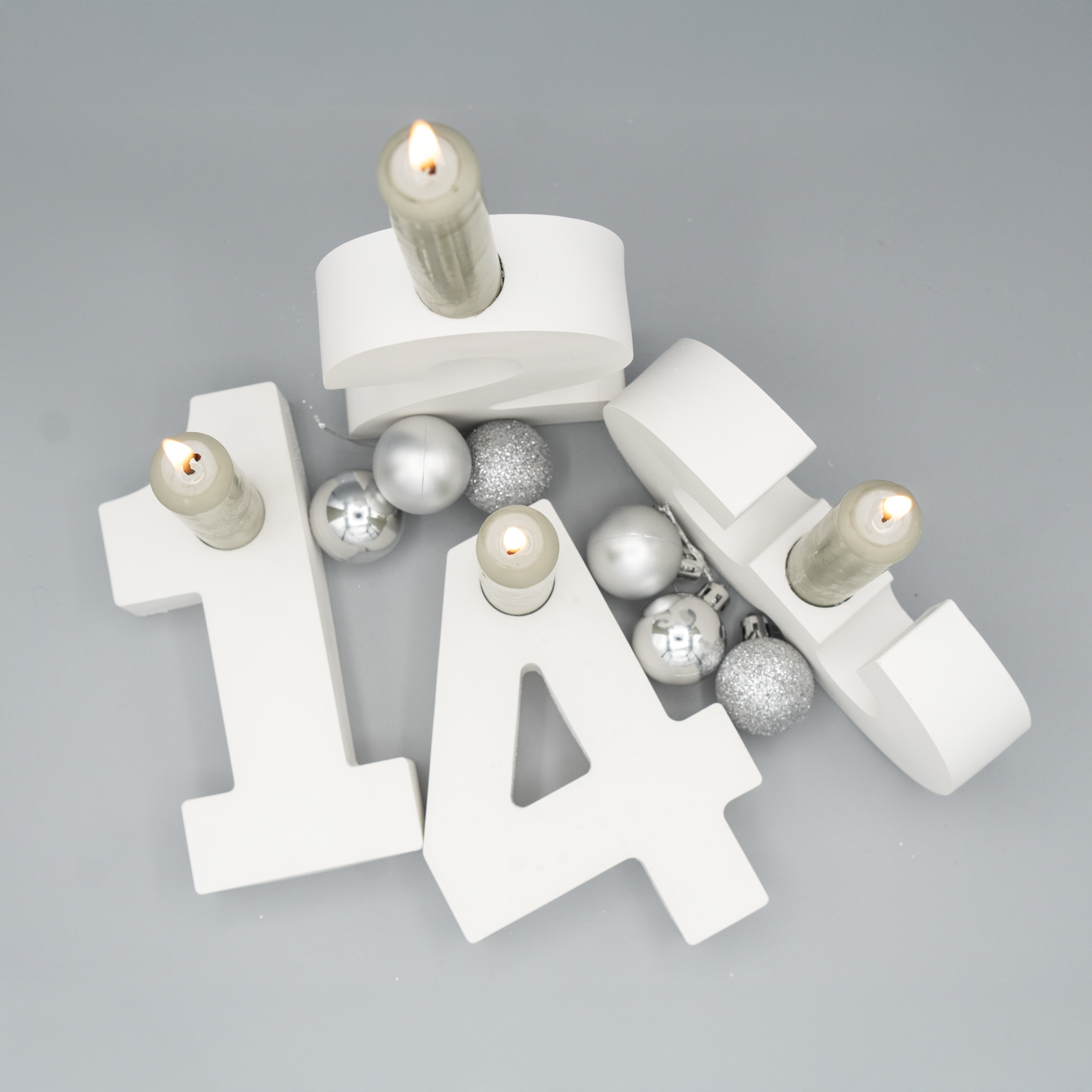 Vier weiße Kerzenhalter in Form der Zahlen 1, 2, 3 und 4 sind aus der Vogelperspektive fotografiert. In jeder Zahl steckt eine brennende Stabkerze. Zwischen den Zahlen liegen silberne und glitzernde Weihnachtskugeln.