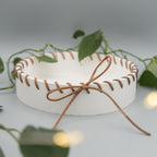 Runde weiße Schale, am Rand dekorativ mit braunem Kunstlederband eingefädelt und einer Schleife gebunden. Im Hintergrund und Vordergrund liegen grüne Philodendron-Blätter.