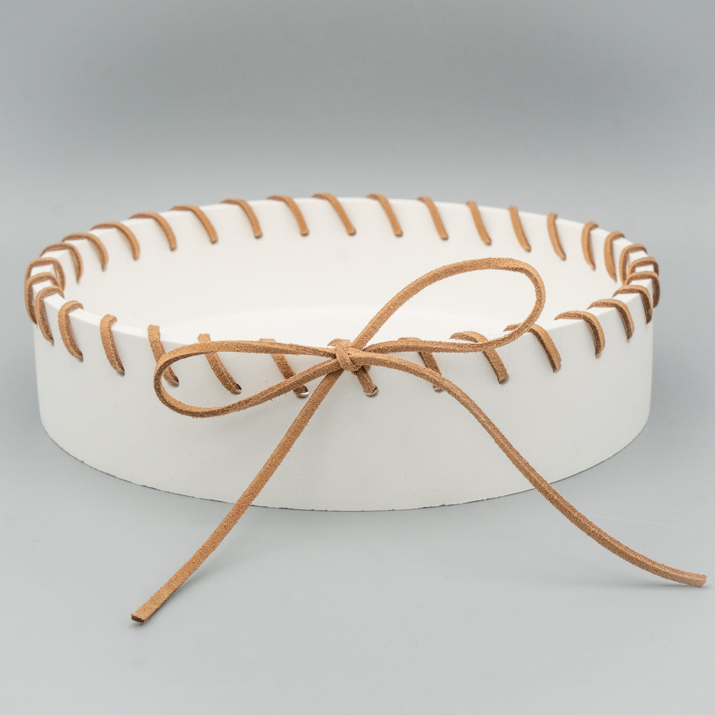 Runde, weiße Schale mit breitem Rand, durch den rundherum ein braunes Kunstlederband gefädelt und vorne zu einer großen Schleife gebunden ist. Die Schale steht auf einem hellen Hintergrund.
