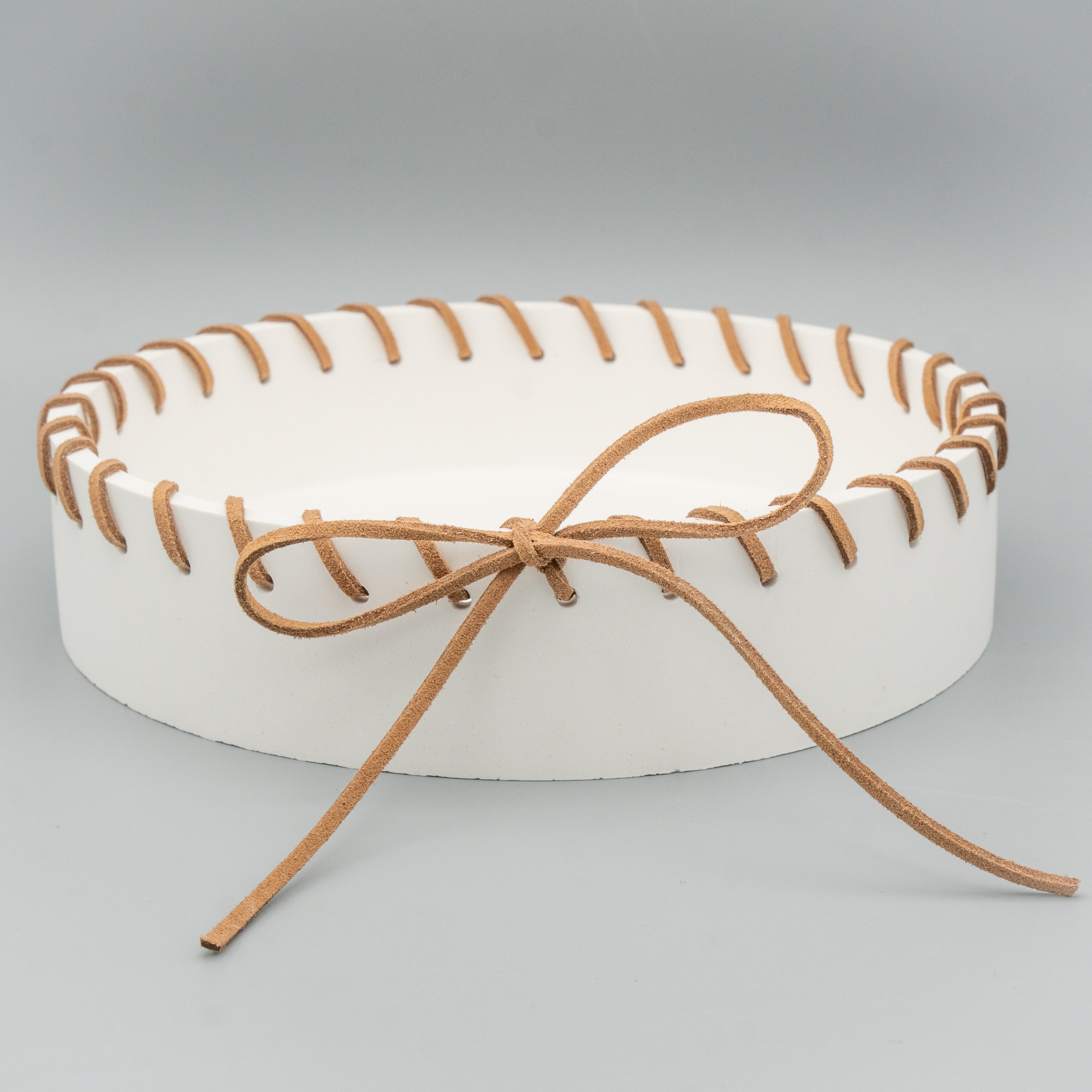 Runde, weiße Schale mit breitem Rand, durch den rundherum ein braunes Kunstlederband gefädelt und vorne zu einer großen Schleife gebunden ist. Die Schale steht auf einem hellen Hintergrund.