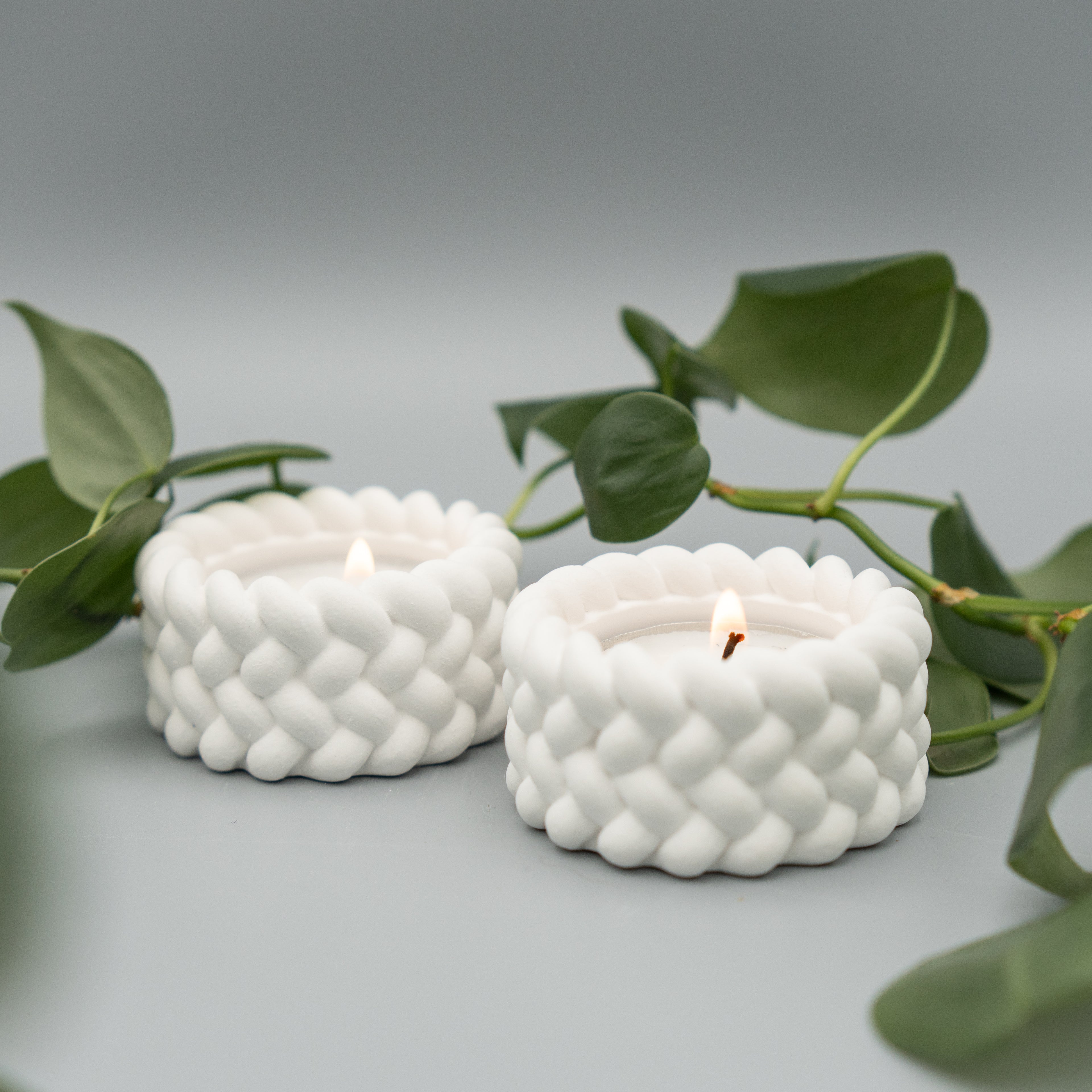 Zwei weiße Teelichthalter mit geflochten wirkender Oberfläche, jeweils mit brennendem Teelicht, stehen nebeneinander auf hellem Untergrund. Im Hintergrund schlängeln sich grüne Philodendron-Ranken.