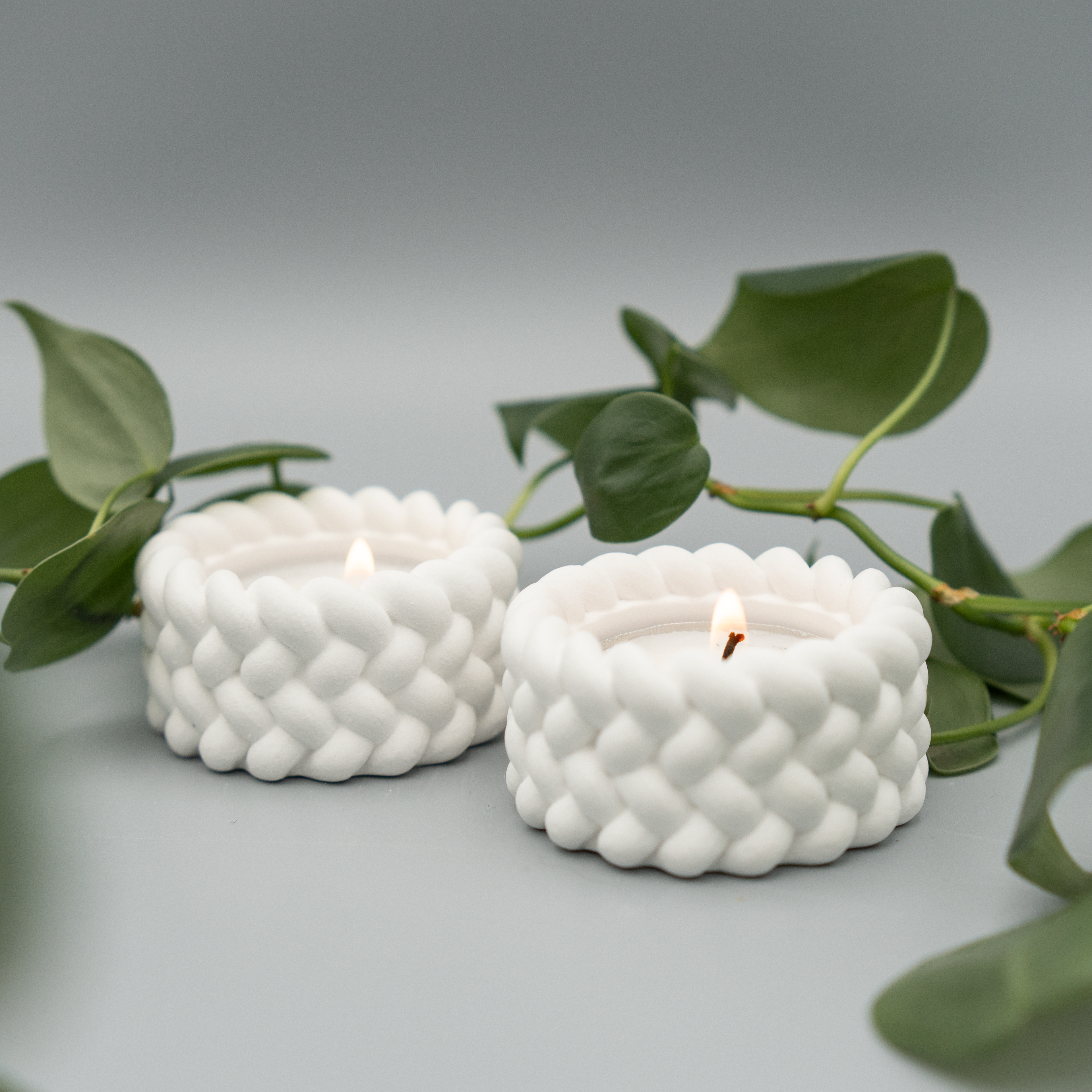 Zwei weiße Teelichthalter mit geflochten wirkender Oberfläche, jeweils mit brennendem Teelicht, stehen nebeneinander auf hellem Untergrund. Im Hintergrund schlängeln sich grüne Philodendron-Ranken.