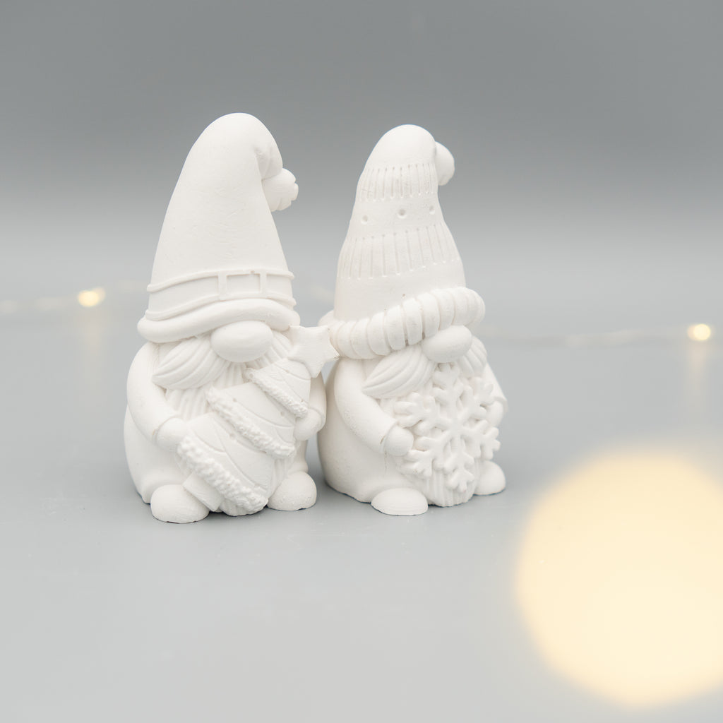 Zwei weiße Weihnachtswichtel-Figuren aus Gießpulver stehen nebeneinander auf einem hellen Untergrund. Der linke Wichtel hält einen Tannenbaum, der rechte eine große Schneeflocke. Im Hintergrund sind zarte Lichtpunkte zu sehen.