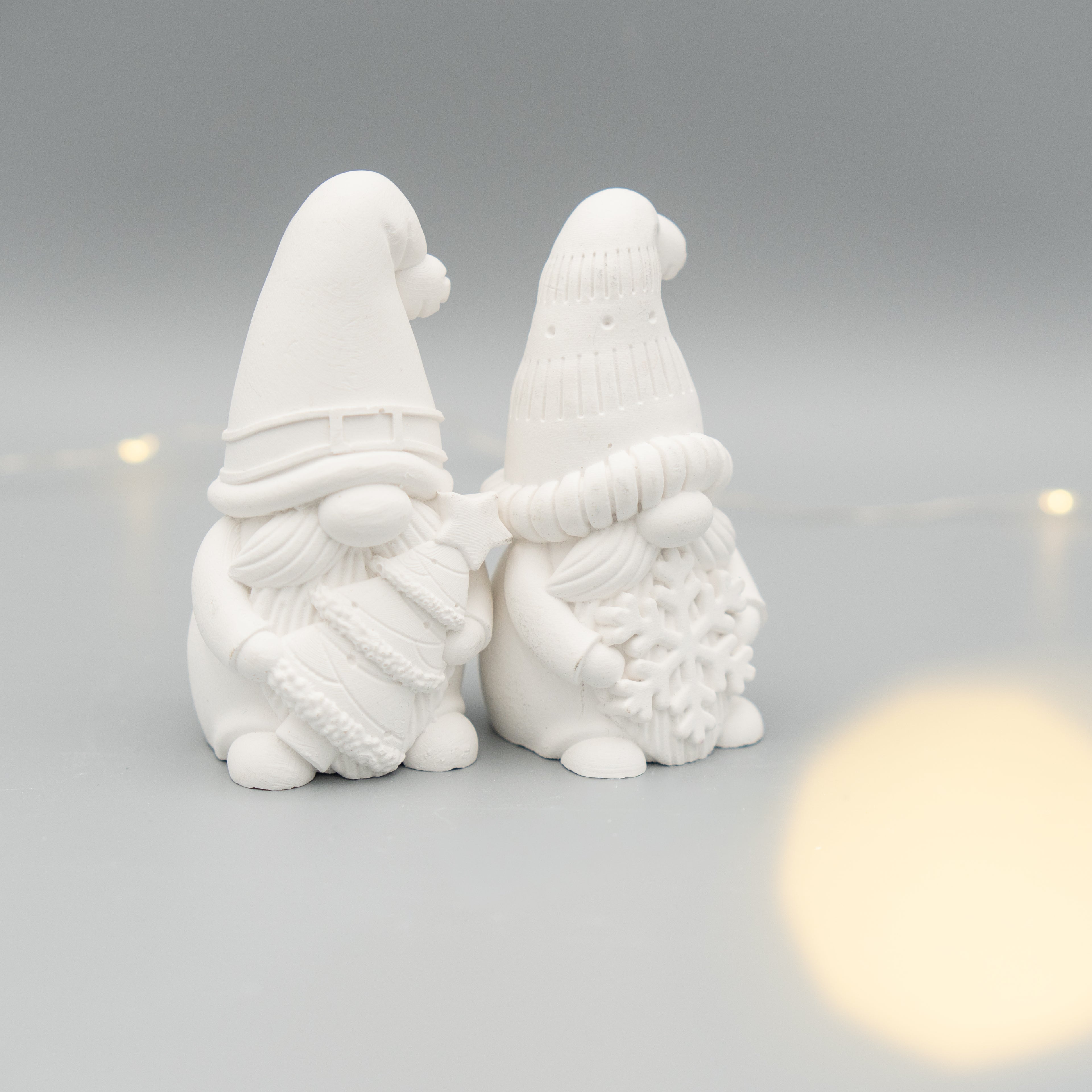 Zwei weiße Weihnachtswichtel-Figuren aus Gießpulver stehen nebeneinander auf einem hellen Untergrund. Der linke Wichtel hält einen Tannenbaum, der rechte eine große Schneeflocke. Im Hintergrund sind zarte Lichtpunkte zu sehen.