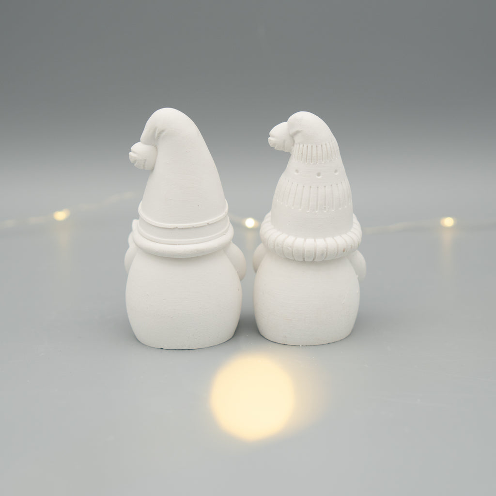 Zwei weiße Wichtelfiguren aus Gießpulver stehen nebeneinander mit dem Rücken zur Kamera. Die Figuren tragen verschiedene Zipfelmützen. Im Hintergrund sorgt eine dezente Lichterkette für stimmungsvolles Licht.