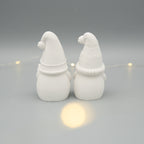 Zwei weiße Wichtelfiguren aus Gießpulver stehen nebeneinander mit dem Rücken zur Kamera. Die Figuren tragen verschiedene Zipfelmützen. Im Hintergrund sorgt eine dezente Lichterkette für stimmungsvolles Licht.