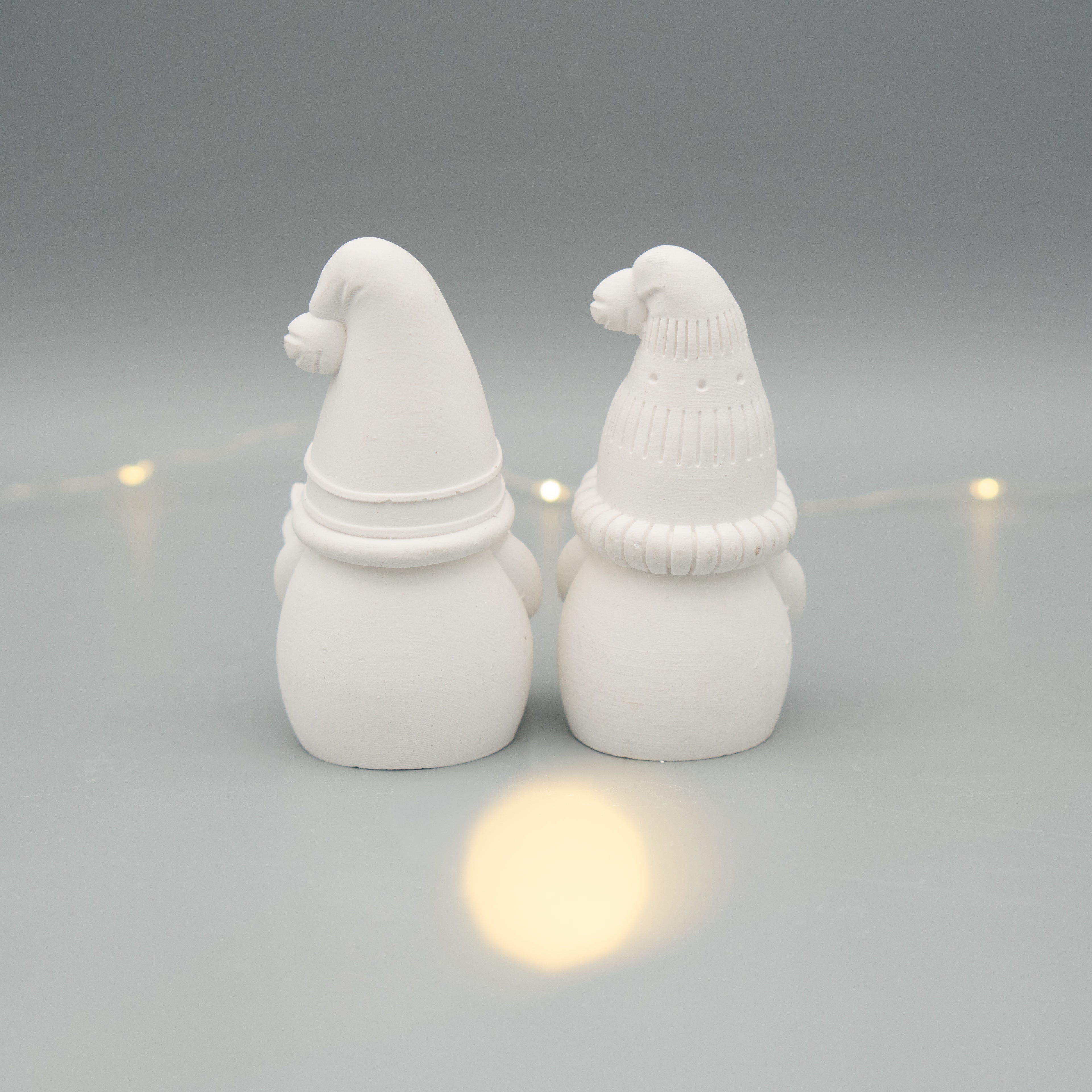 Zwei weiße Wichtelfiguren aus Gießpulver stehen nebeneinander mit dem Rücken zur Kamera. Die Figuren tragen verschiedene Zipfelmützen. Im Hintergrund sorgt eine dezente Lichterkette für stimmungsvolles Licht.