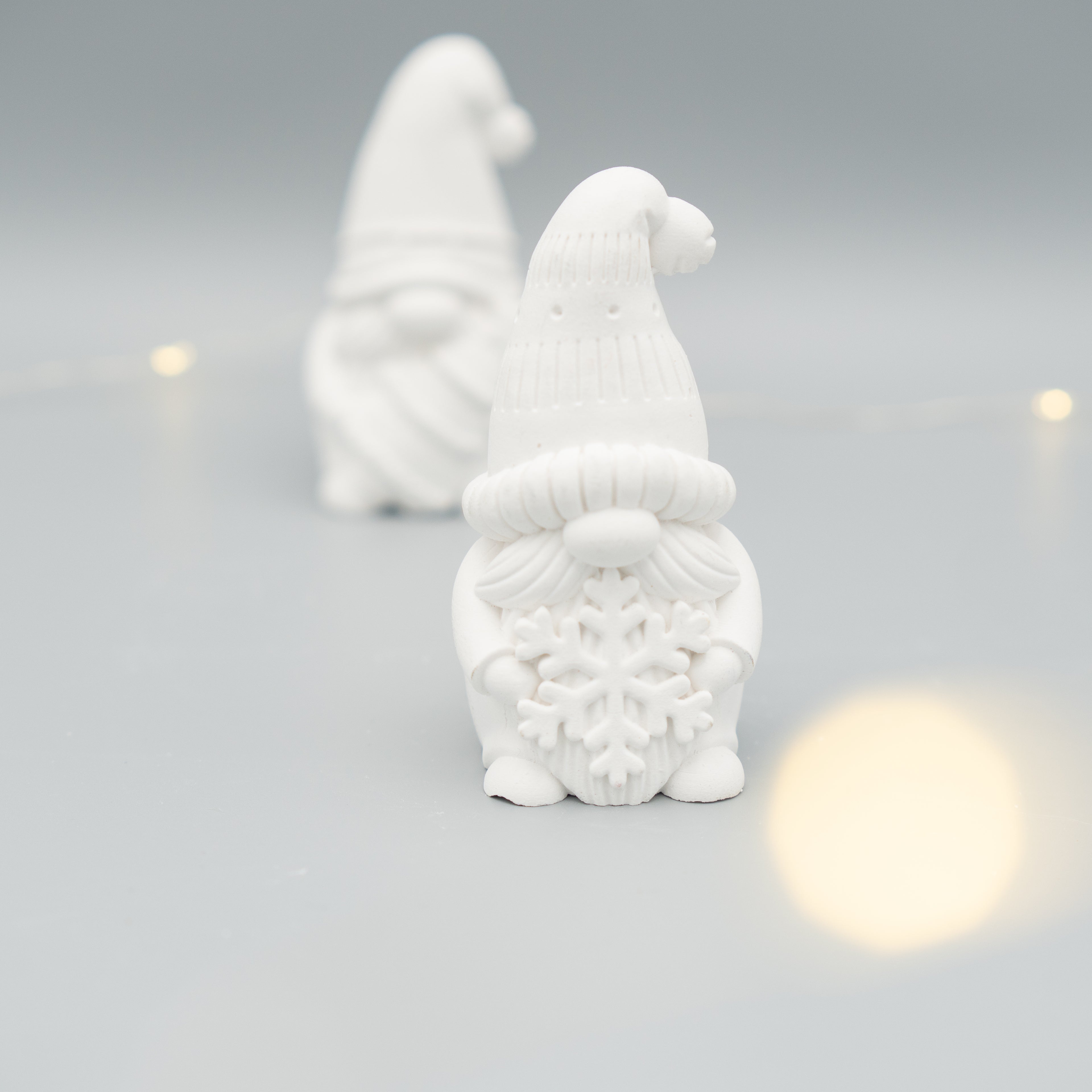 Im Vordergrund steht eine weiße Wichtelfigur aus Gießpulver mit einer großen Schneeflocke in den Händen. Im Hintergrund ist eine weitere, unscharfe Wichtelfigur zu sehen. Der Hintergrund ist hell, einzelne Lichter sorgen für eine sanfte Atmosphäre.