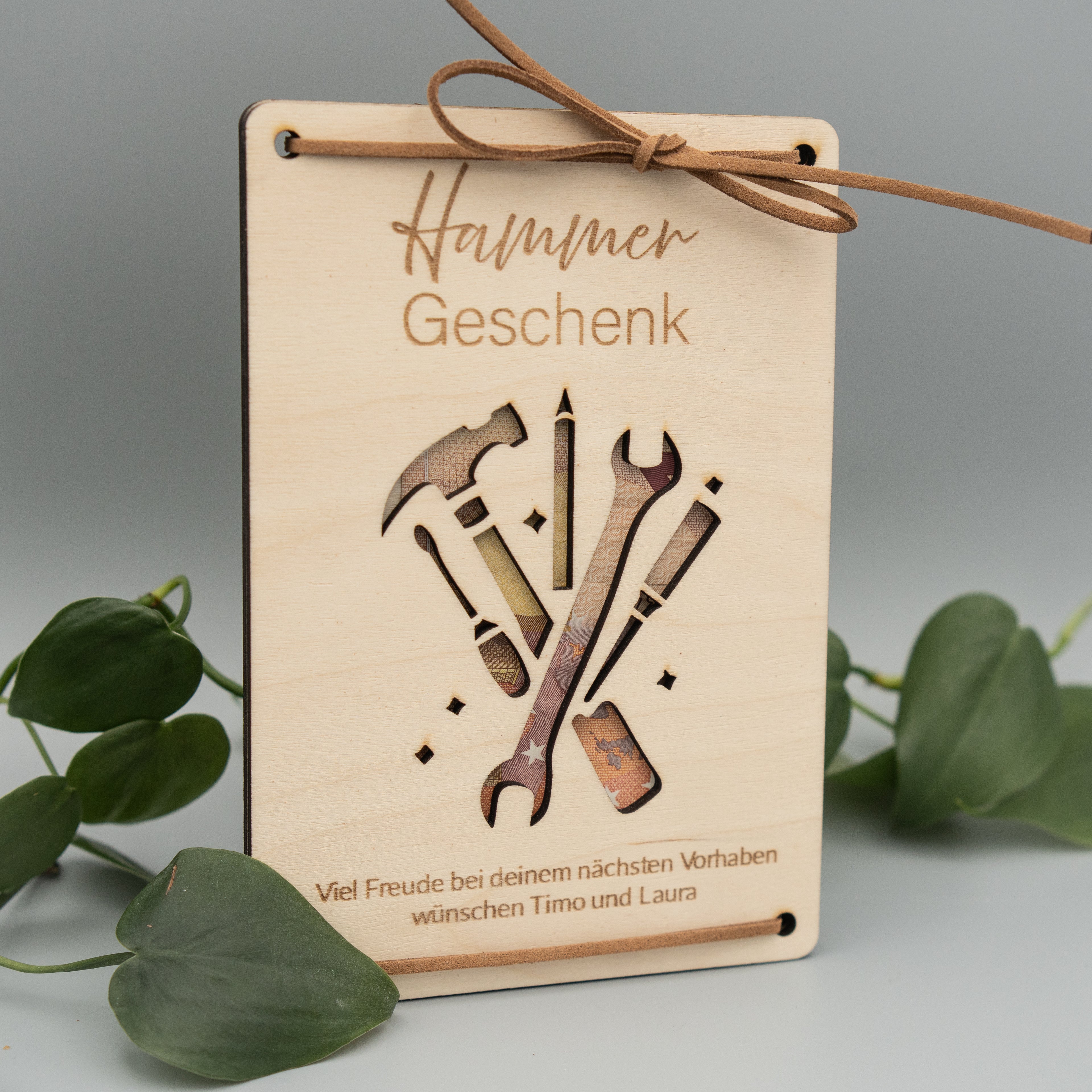 Rechteckige Holztafel mit graviertem Schriftzug ‘Hammer Geschenk’ und einem ausgeschnittenen Motiv mit Werkzeugen wie Hammer, Schraubendreher und Schraubenschlüssel. Unten steht die Gravur: ‘Viel Freude bei deinem nächsten Vorhaben wünschen Timo und Laura’. Die Tafel ist mit braunen Kunstlederbändern dekorativ zusammengebunden, umgeben von grünen Philodendron-Blättern.