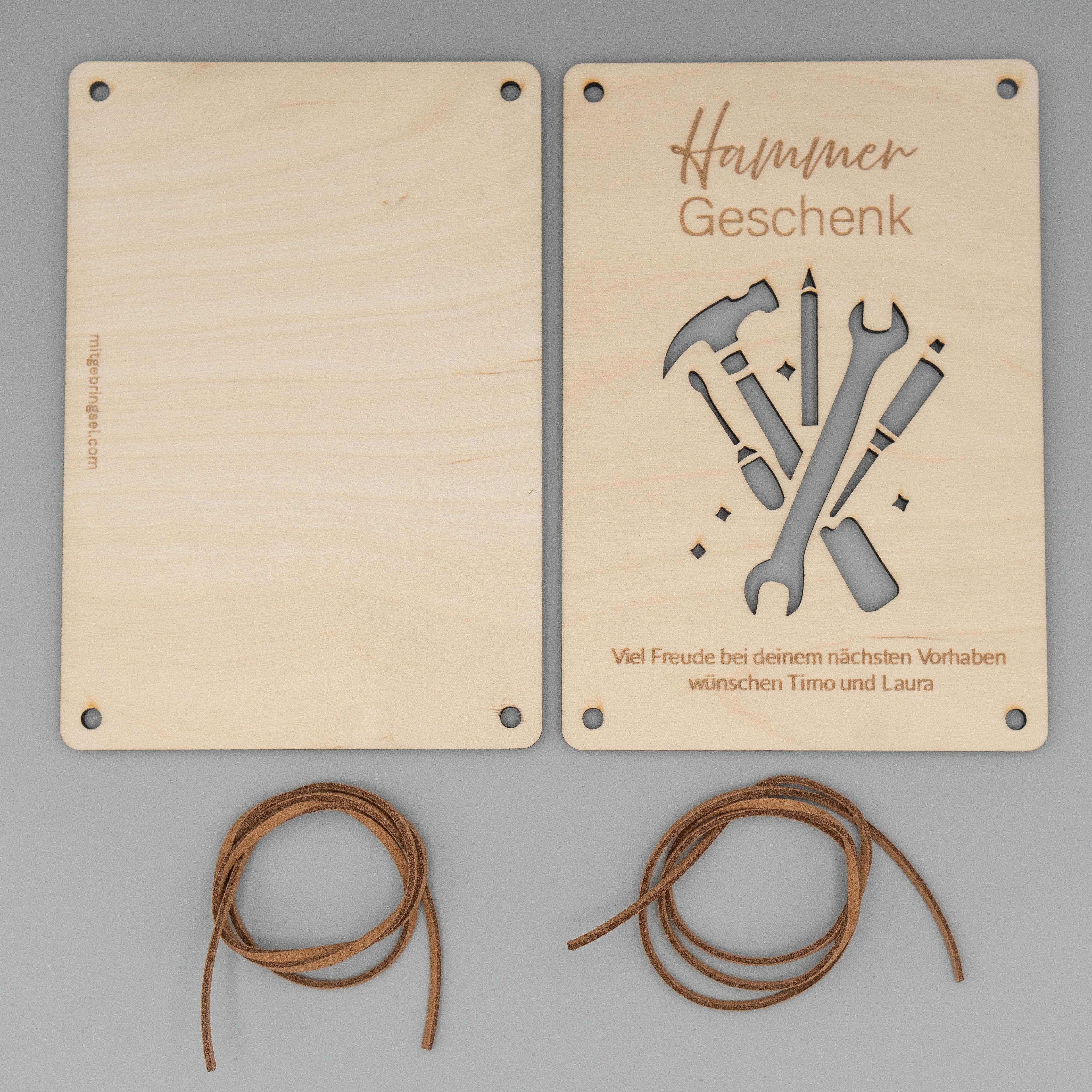 Zwei rechteckige Holzplatten liegen nebeneinander auf einem grauen Hintergrund. Die rechte Platte ist graviert mit ‘Hammer Geschenk’, zeigt ein ausgeschnittenes Werkzeug-Motiv und trägt unten die Gravur ‘Viel Freude bei deinem nächsten Vorhaben wünschen Timo und Laura’. Darunter sind zwei braune Kunstlederbänder zu sehen.