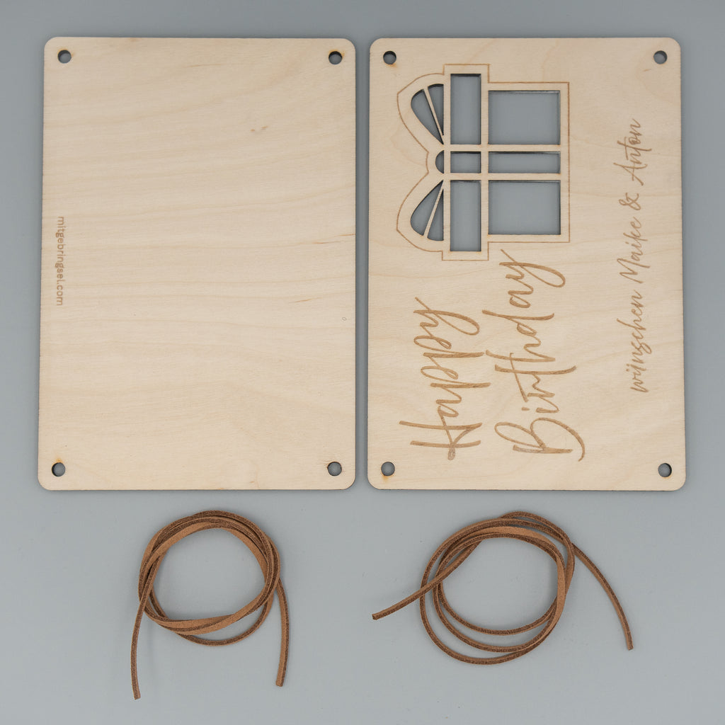 Zwei Holzplatten liegen nebeneinander, eine mit einer Gravur ‘Happy Birthday’ und einem ausgeschnittenen Geschenk-Motiv sowie dem Schriftzug ‘wünschen Maike & Anton’, die andere ohne Verzierung. Darunter befinden sich zwei braune Kunstlederschnüre.