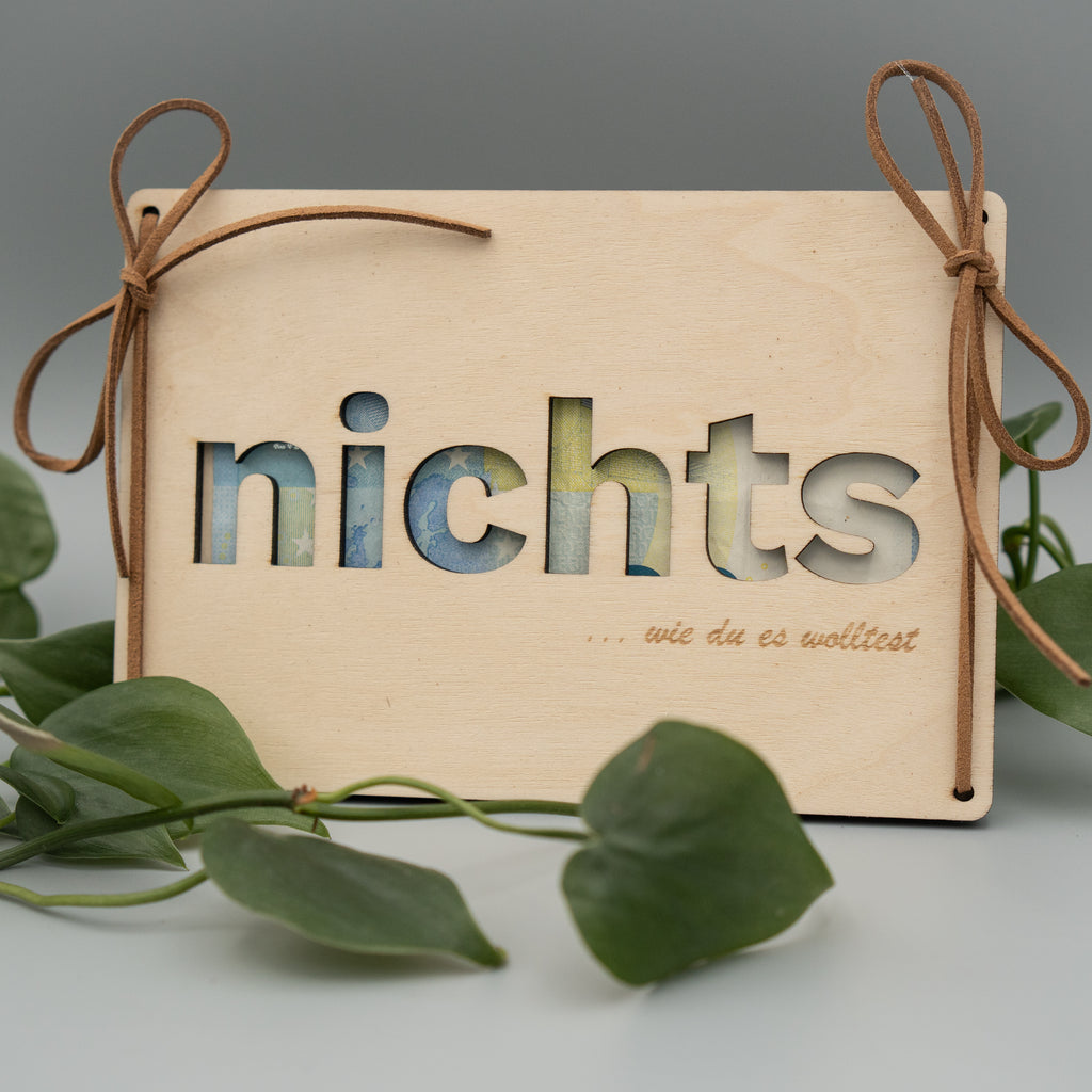 Holztafel mit ausgeschnittenem Wort ‘nichts’, dahinter sind Geldscheine sichtbar. Links und rechts sind Kunstlederschnüre zu Schleifen gebunden, unten ist ein Schriftzug mit ‘… wie du es wolltest’ eingraviert. Im Vordergrund liegen grüne Philodendron-Blätter auf hellem Untergrund.