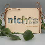 Holztafel mit ausgeschnittenem Wort ‘nichts’, dahinter sind Geldscheine sichtbar. Links und rechts sind Kunstlederschnüre zu Schleifen gebunden, unten ist ein Schriftzug mit ‘… wie du es wolltest’ eingraviert. Im Vordergrund liegen grüne Philodendron-Blätter auf hellem Untergrund.