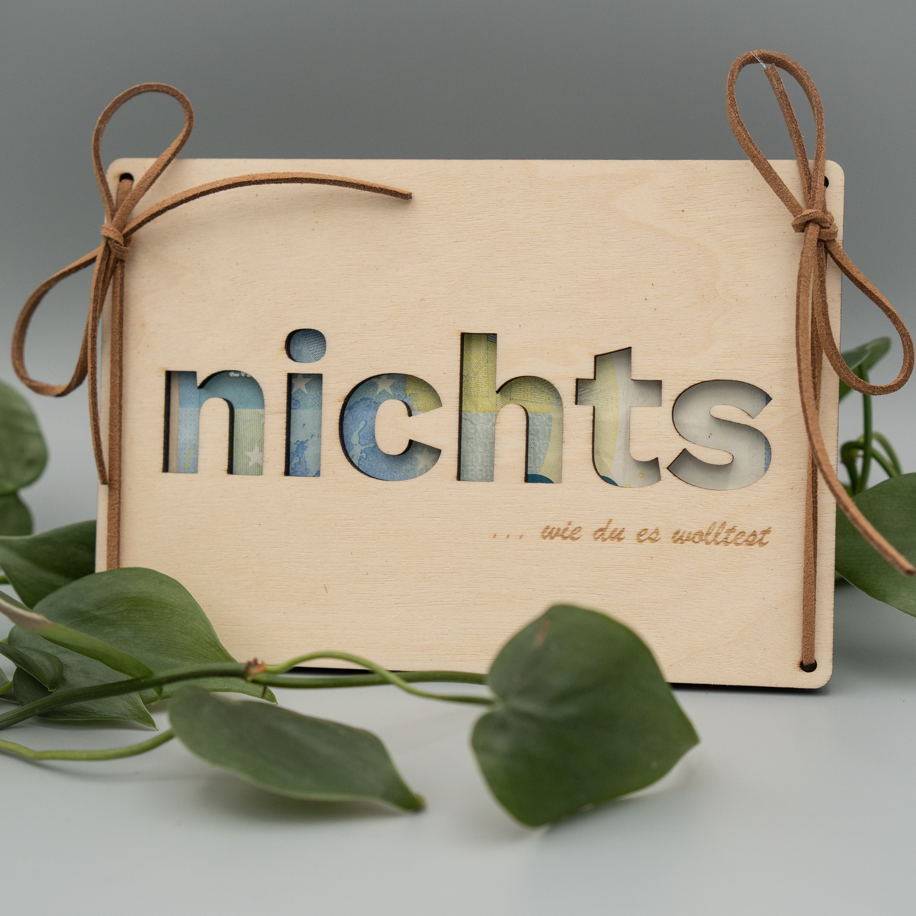 Holztafel mit ausgeschnittenem Wort ‘nichts’, dahinter sind Geldscheine sichtbar. Links und rechts sind Kunstlederschnüre zu Schleifen gebunden, unten ist ein Schriftzug mit ‘… wie du es wolltest’ eingraviert. Im Vordergrund liegen grüne Philodendron-Blätter auf hellem Untergrund.