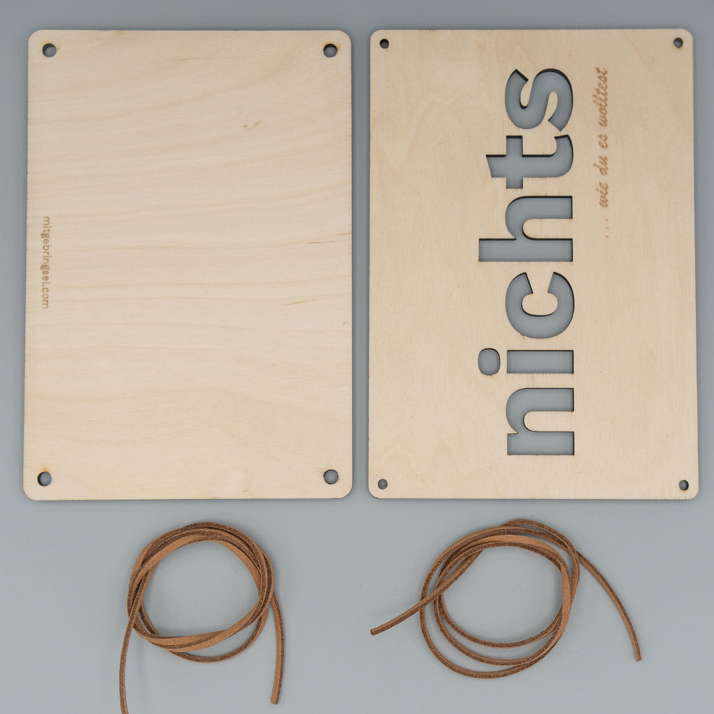 Zwei rechteckige Holzplatten liegen nebeneinander, eine davon ist leer, die andere zeigt vertikal das ausgeschnittene Wort ‘nichts’ sowie die Gravur ‘… wie du es wolltest’. Darunter befinden sich zwei zusammengerollte braune Kunstlederbänder.