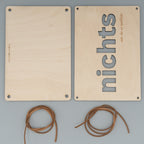 Zwei rechteckige Holzplatten liegen nebeneinander, eine davon ist leer, die andere zeigt vertikal das ausgeschnittene Wort ‘nichts’ sowie die Gravur ‘… wie du es wolltest’. Darunter befinden sich zwei zusammengerollte braune Kunstlederbänder.