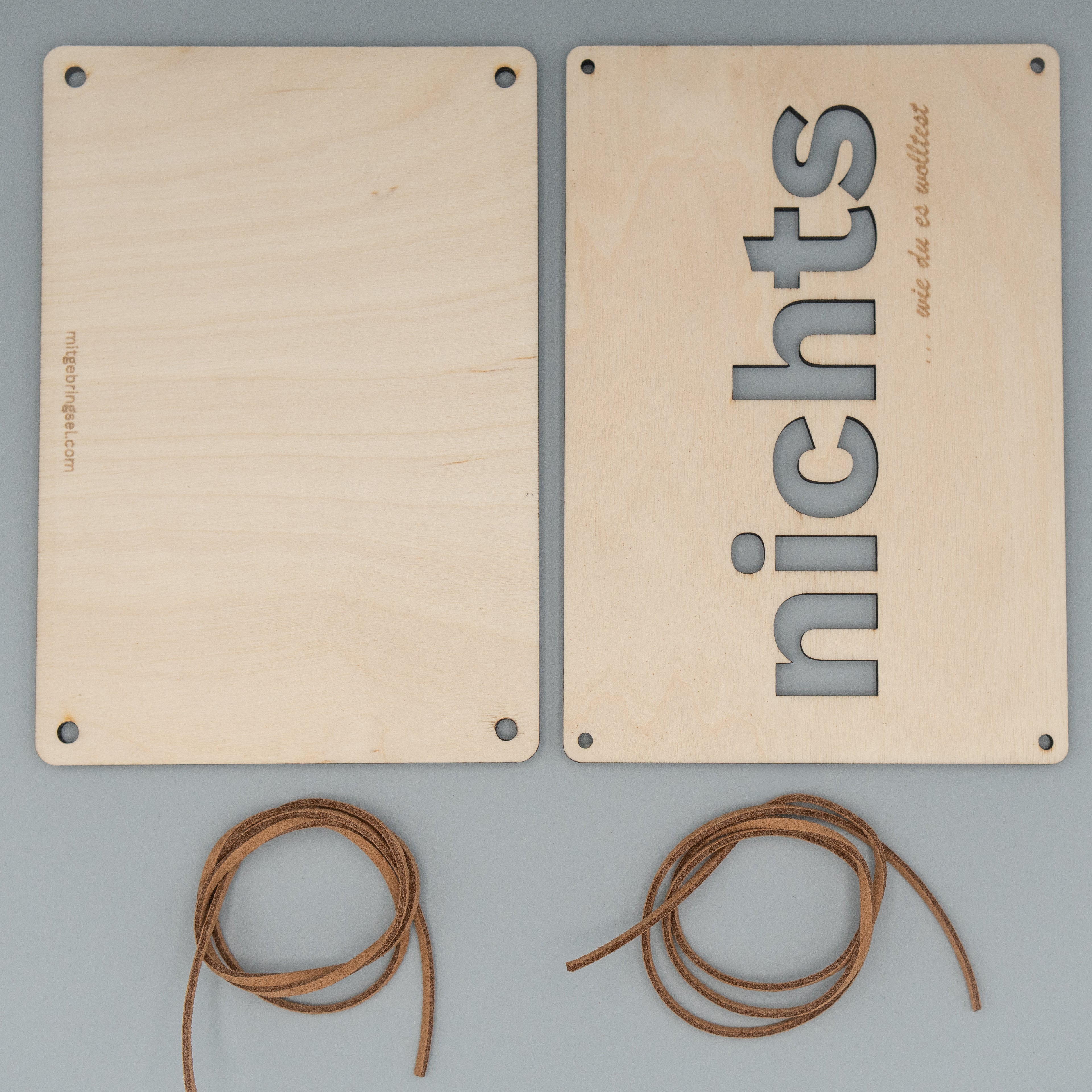 Zwei rechteckige Holzplatten liegen nebeneinander, eine davon ist leer, die andere zeigt vertikal das ausgeschnittene Wort ‘nichts’ sowie die Gravur ‘… wie du es wolltest’. Darunter befinden sich zwei zusammengerollte braune Kunstlederbänder.