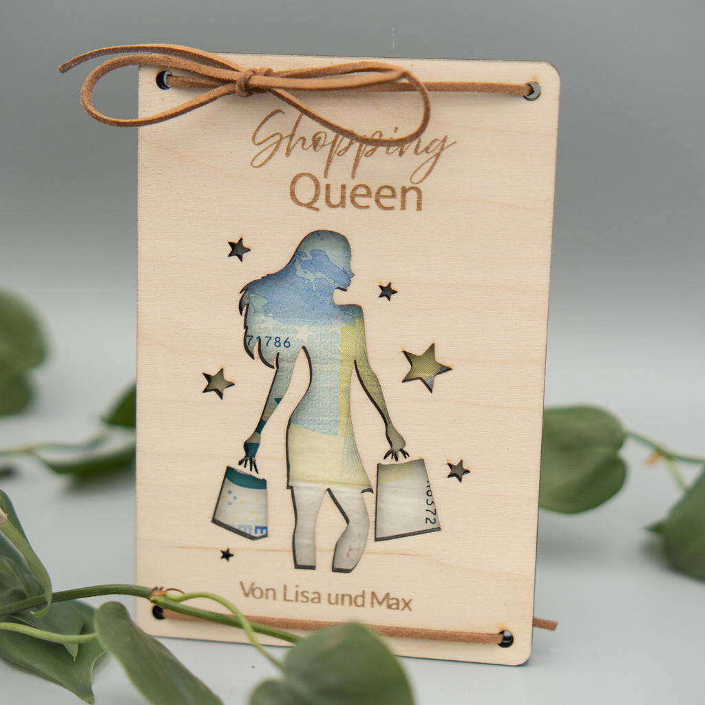 Rechteckige Holztafel mit graviertem Schriftzug ‘Shopping Queen’ und einer ausgeschnittenen Silhouette einer Frau, die Einkaufstüten trägt. Die Tafel ist mit einem braunen Kunstlederband dekorativ gebunden. Unten steht der Text ‘Von Lisa und Max’. Im Vordergrund liegen grüne Philodendron-Blätter.