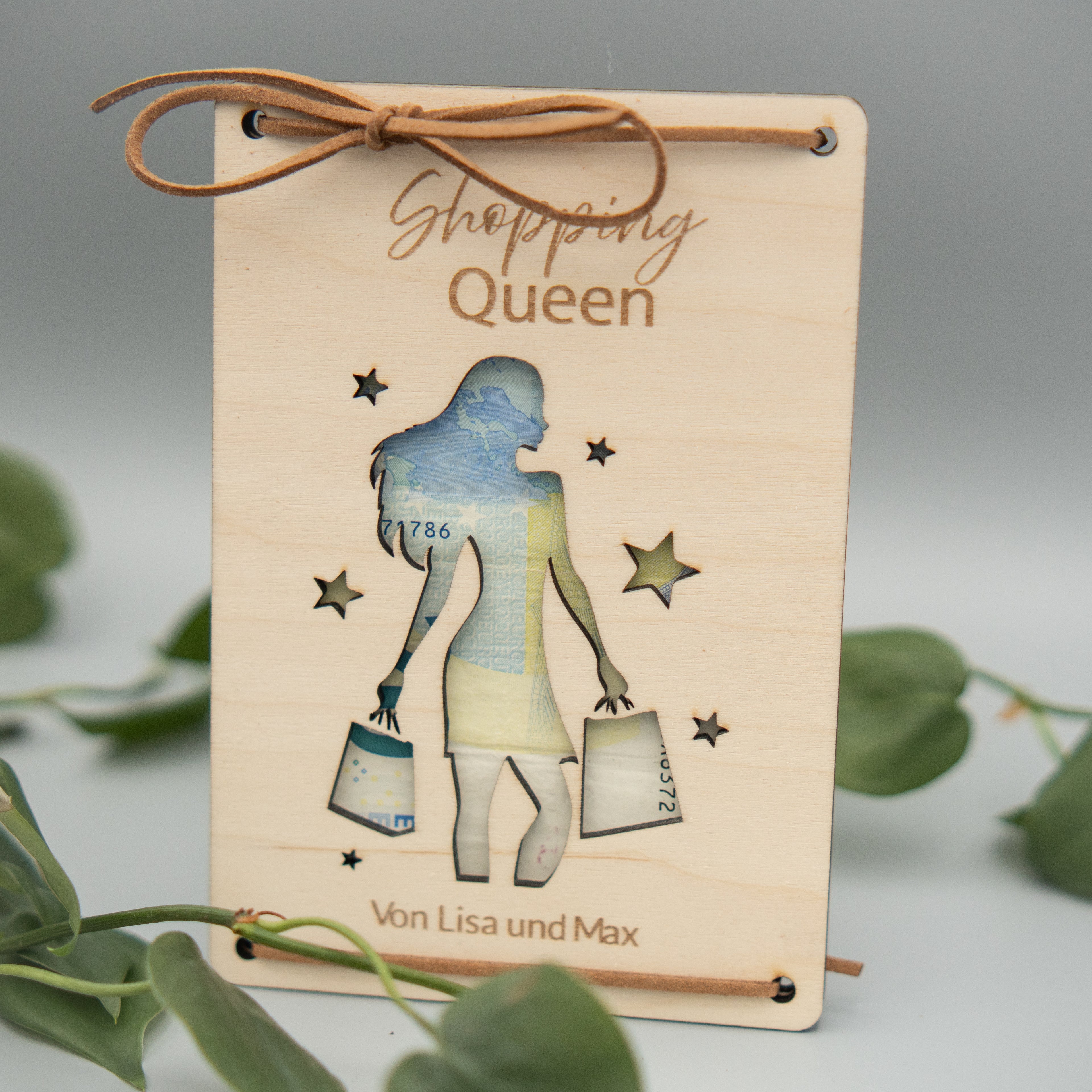 Rechteckige Holztafel mit graviertem Schriftzug ‘Shopping Queen’ und einer ausgeschnittenen Silhouette einer Frau, die Einkaufstüten trägt. Die Tafel ist mit einem braunen Kunstlederband dekorativ gebunden. Unten steht der Text ‘Von Lisa und Max’. Im Vordergrund liegen grüne Philodendron-Blätter.