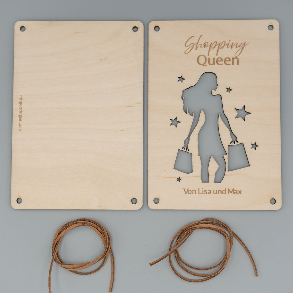 Zwei Holzplatten liegen nebeneinander, die rechte zeigt eine Gravur ‘Shopping Queen’ und ein ausgestanztes Motiv einer Frau mit Einkaufstaschen sowie mehreren Sternen. Unten ist der Schriftzug ‘Von Lisa und Max’ eingraviert. Darunter liegen zwei zusammengerollte braune Kunstlederbänder.