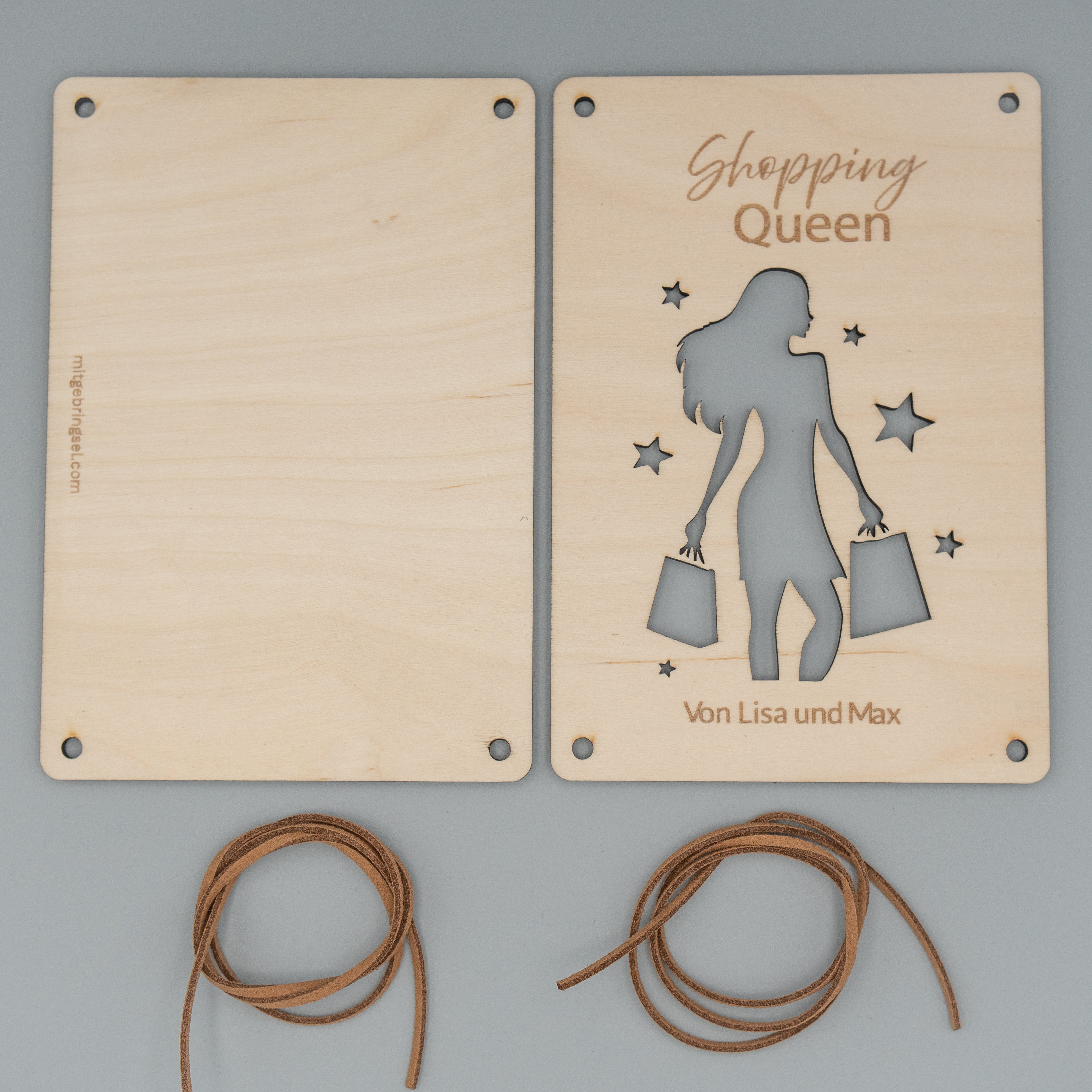 Zwei Holzplatten liegen nebeneinander, die rechte zeigt eine Gravur ‘Shopping Queen’ und ein ausgestanztes Motiv einer Frau mit Einkaufstaschen sowie mehreren Sternen. Unten ist der Schriftzug ‘Von Lisa und Max’ eingraviert. Darunter liegen zwei zusammengerollte braune Kunstlederbänder.