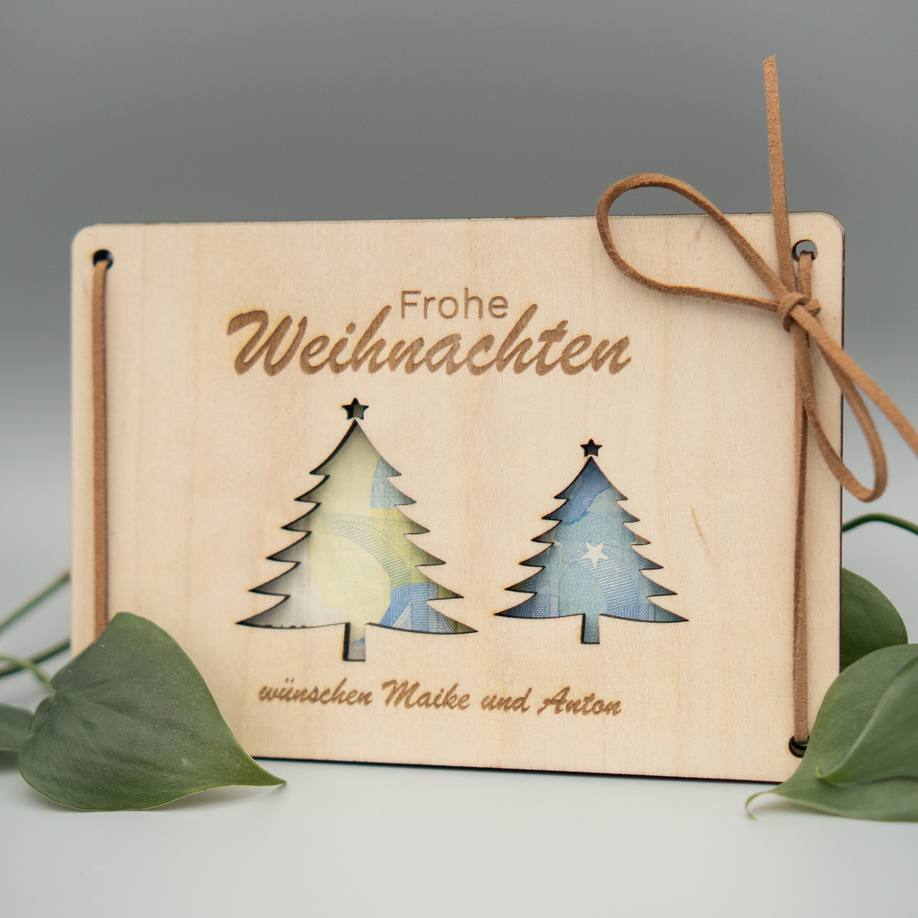 Rechteckige Holztafel mit der Gravur ‘Frohe Weihnachten’ oben und zwei ausgeschnittenen Weihnachtsbaum-Silhouetten. Die Tafel ist mit braunen Kunstlederbändern dekorativ gebunden. Unten steht der Schriftzug ‘wünschen Maike und Anton’. Die Tafel ist umgeben von grünen Philodendron-Blättern.