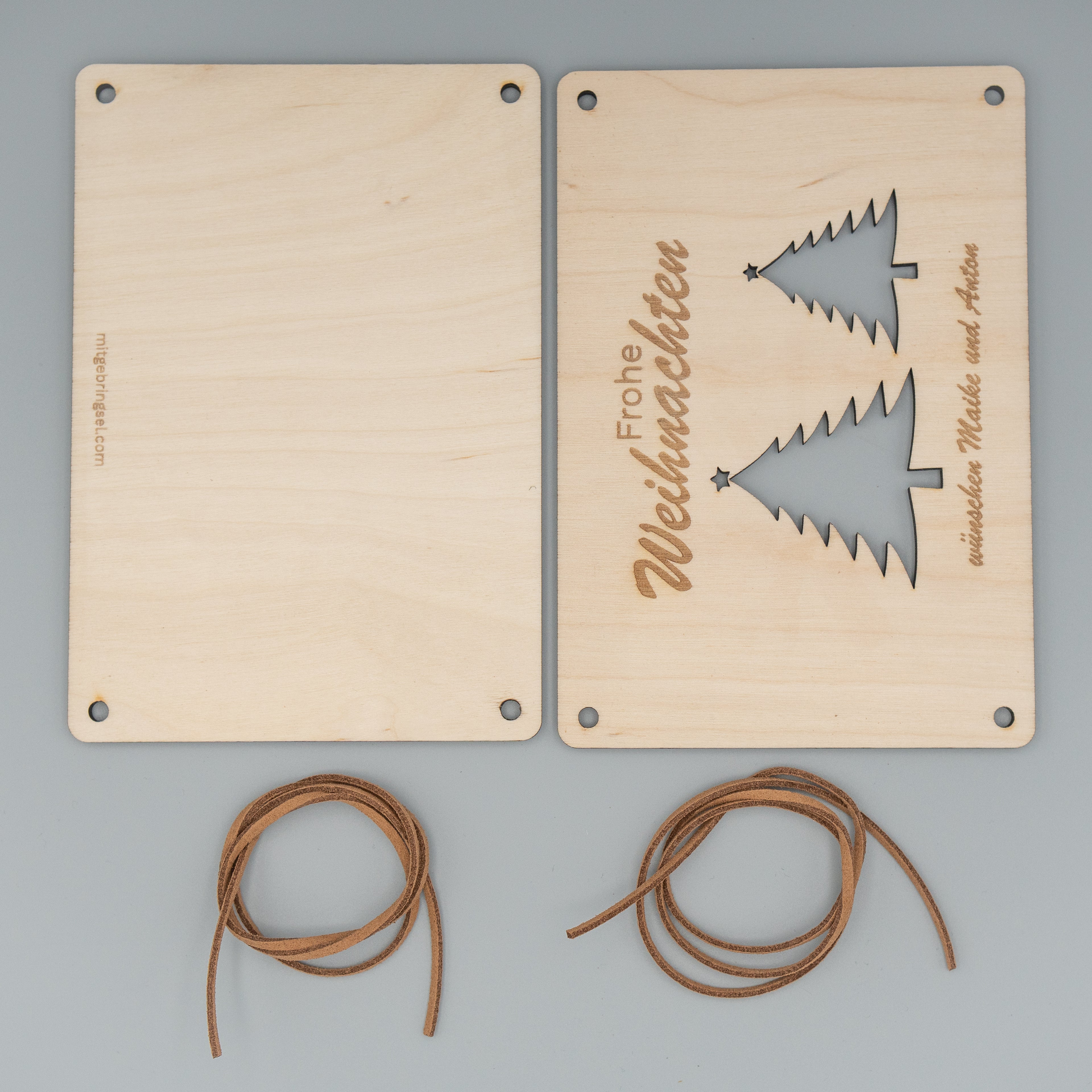 Zwei rechteckige Holzplatten liegen nebeneinander, die rechte zeigt vertikal die Gravur ‘Frohe Weihnachten’ und zwei ausgestanzte Weihnachtsbaum-Silhouetten. Am unteren Rand steht ‘wünschen Maike und Anton’. Darunter liegen zwei zusammengerollte braune Kunstlederbänder.