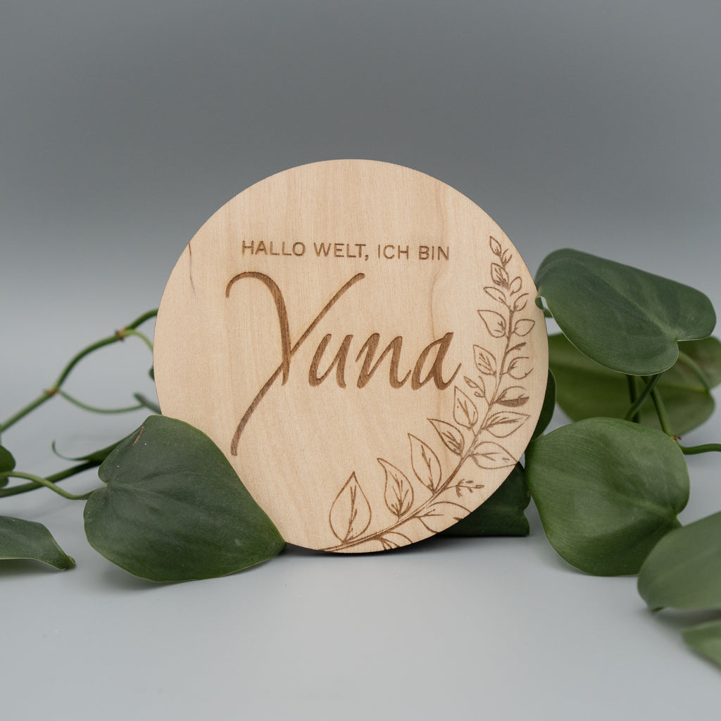 Runde Holzscheibe mit Gravur ‘Hallo Welt, ich bin Yuna’ und einem filigranen Blattdekor, dekorativ von grünen Philodendron-Blättern umrahmt, auf hellem Hintergrund arrangiert.