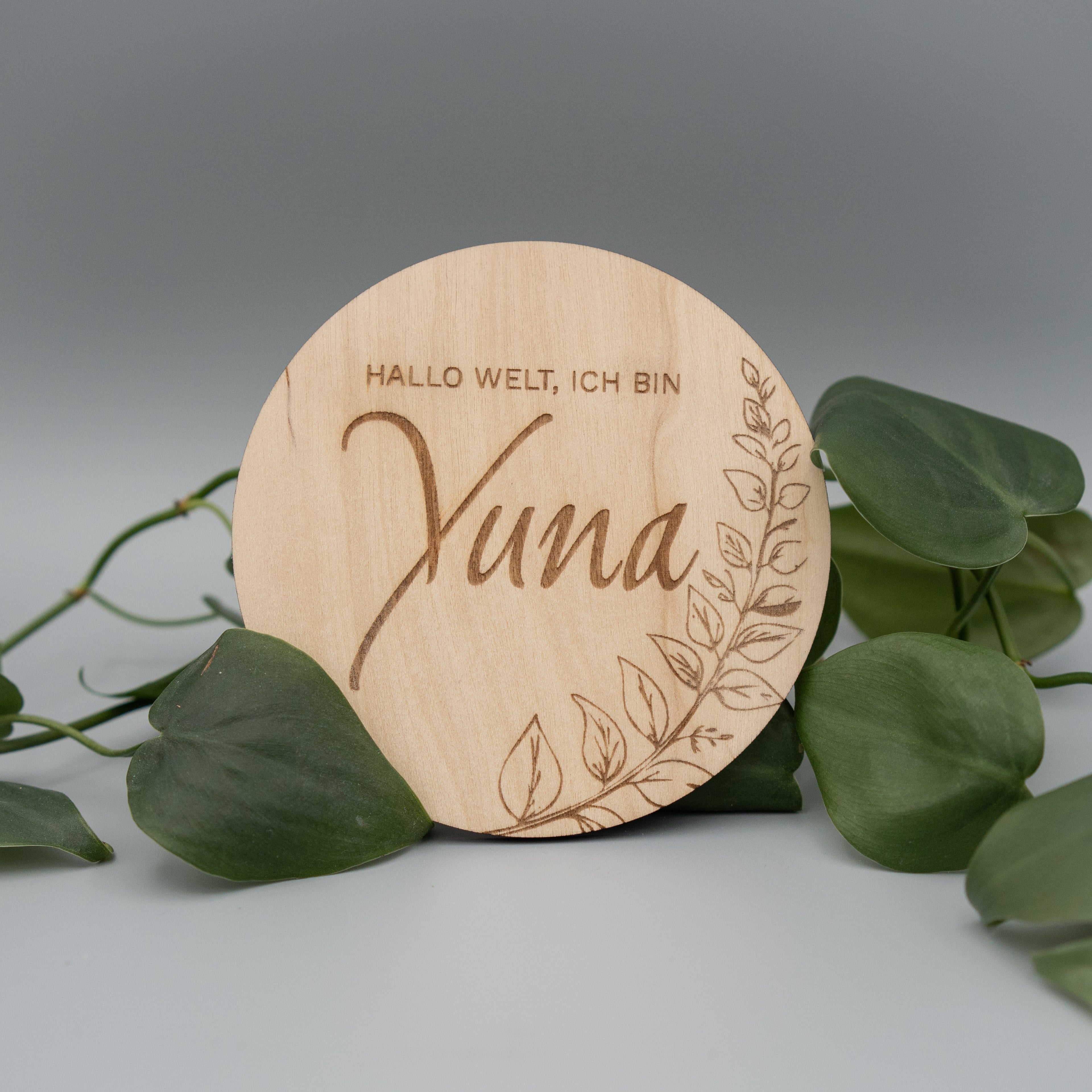 Runde Holzscheibe mit Gravur ‘Hallo Welt, ich bin Yuna’ und einem filigranen Blattdekor, dekorativ von grünen Philodendron-Blättern umrahmt, auf hellem Hintergrund arrangiert.