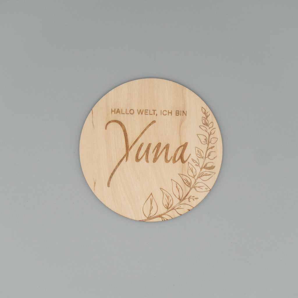 Runde Holzscheibe mit dem eingravierten Text ‘Hallo Welt, ich bin Yuna’ und einem zarten Blattmotiv am Rand, auf grauem Hintergrund fotografiert.