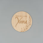 Runde Holzscheibe mit dem eingravierten Text ‘Hallo Welt, ich bin Yuna’ und einem zarten Blattmotiv am Rand, auf grauem Hintergrund fotografiert.