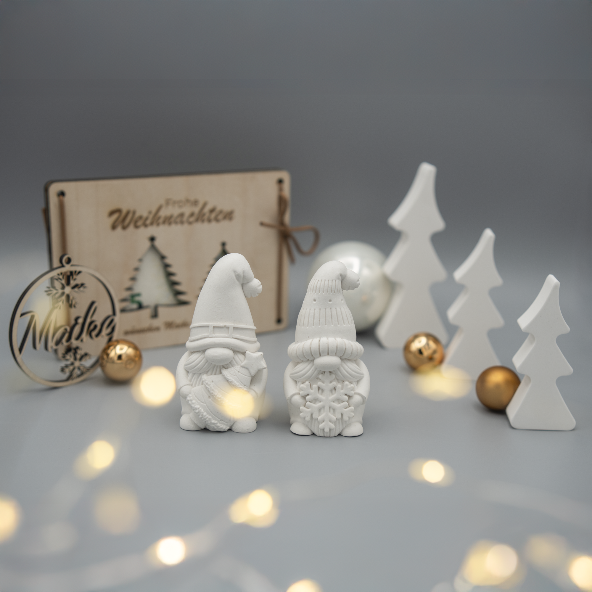 Weihnachtliche Arrangement mit zwei weißen Wichtelfiguren aus Gießpulver, mehreren weißen Weihnachtsbaumfiguren, goldenen und silbernen Kugeln. Im Hintergrund stehen eine gravierte Holzkarte mit Weihnachtsmotiv und ein Holzanhänger mit dem Namen ‘Maike’. Lichterketten sorgen für eine warme Stimmung.