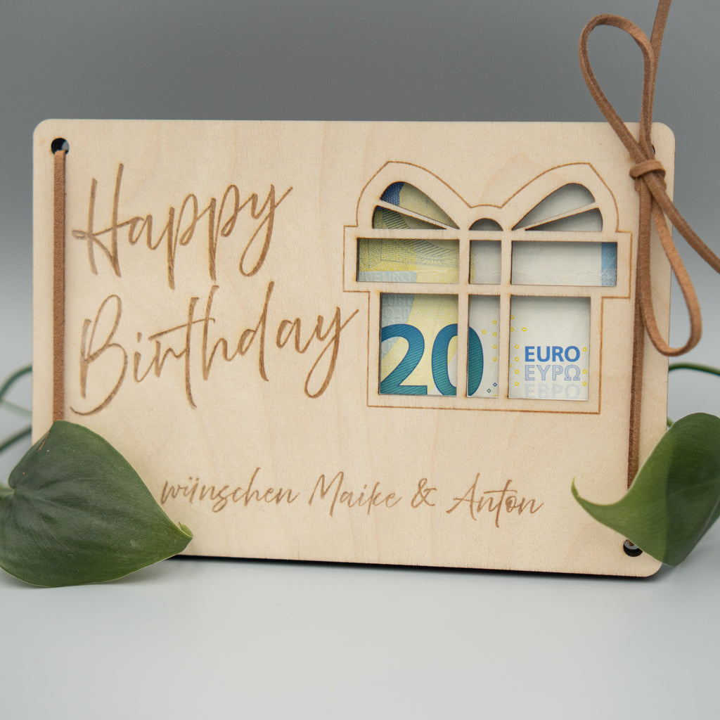 Eine rechteckige Holzkarte mit graviertem Schriftzug „Happy Birthday“ und darunter „wünschen Maike & Anton“ liegt auf einer glatten Oberfläche. Auf der rechten Seite ist ein Geschenkmotiv mit Ausschnitten eingearbeitet, in dem ein 20-Euro-Schein sichtbar platziert ist. Eine Lederschnur ist durch ein Loch in der Ecke gefädelt und locker gebunden. Zwei grüne Blätter dekorieren die Ecken der Karte.